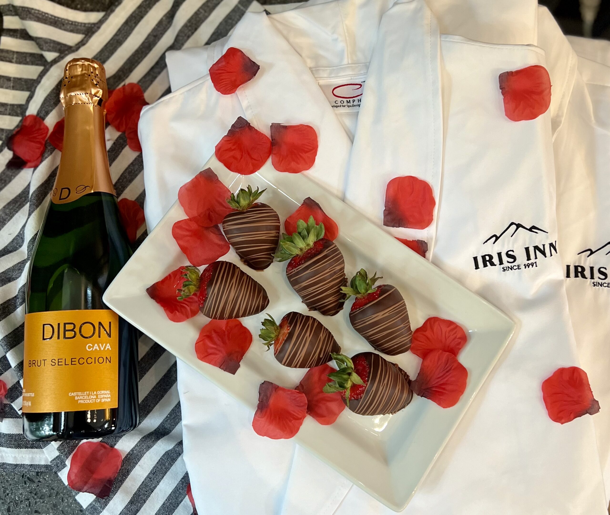 Overhead shot of a romantic welcome package featuring a bottle of Dibon Cava Brut Seleccion sparkling wine and a platter of chocolate-covered strawberries, arranged on white Iris Inn robes and red rose petals.