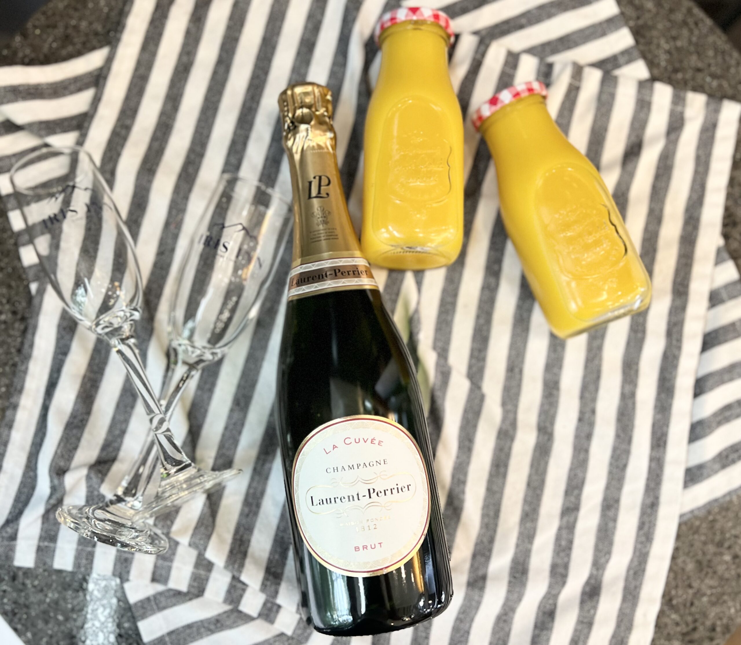 Overhead shot of a mimosa setup featuring a bottle of Laurent-Perrier La Cuvée Champagne, two Iris Inn engraved champagne flutes, and two bottles of orange juice, all on a gray and white striped cloth.