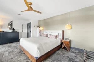 Bedroom with a made bed, nightstand, lamp, ceiling fan, and soft natural light.