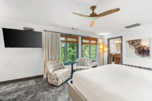 Bedroom with bed, pillows, wall-mounted TV, ceiling fan, and large windows with treatments.