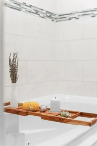 Wooden bath caddy with candle, bath bomb, and lavender on a white bathtub.