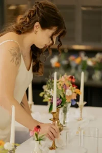 A woman in a white wedding dress stands indoors surrounded by candles and flower arrangements, holding roses.