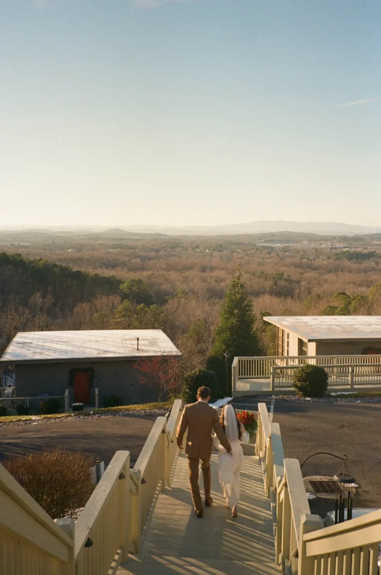 groom walks down outdoor wooden stairs toward a scenic mountain overlook at sunset.