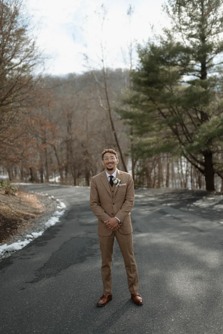 Groom in a tan suit stands smiling on a paved path surrounded by winter trees.