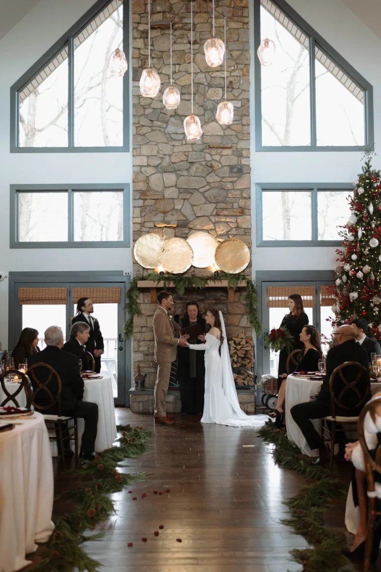 wedding ceremony taking place inside a vaulted room in front of a large stone fireplace and floor-to-ceiling windows.