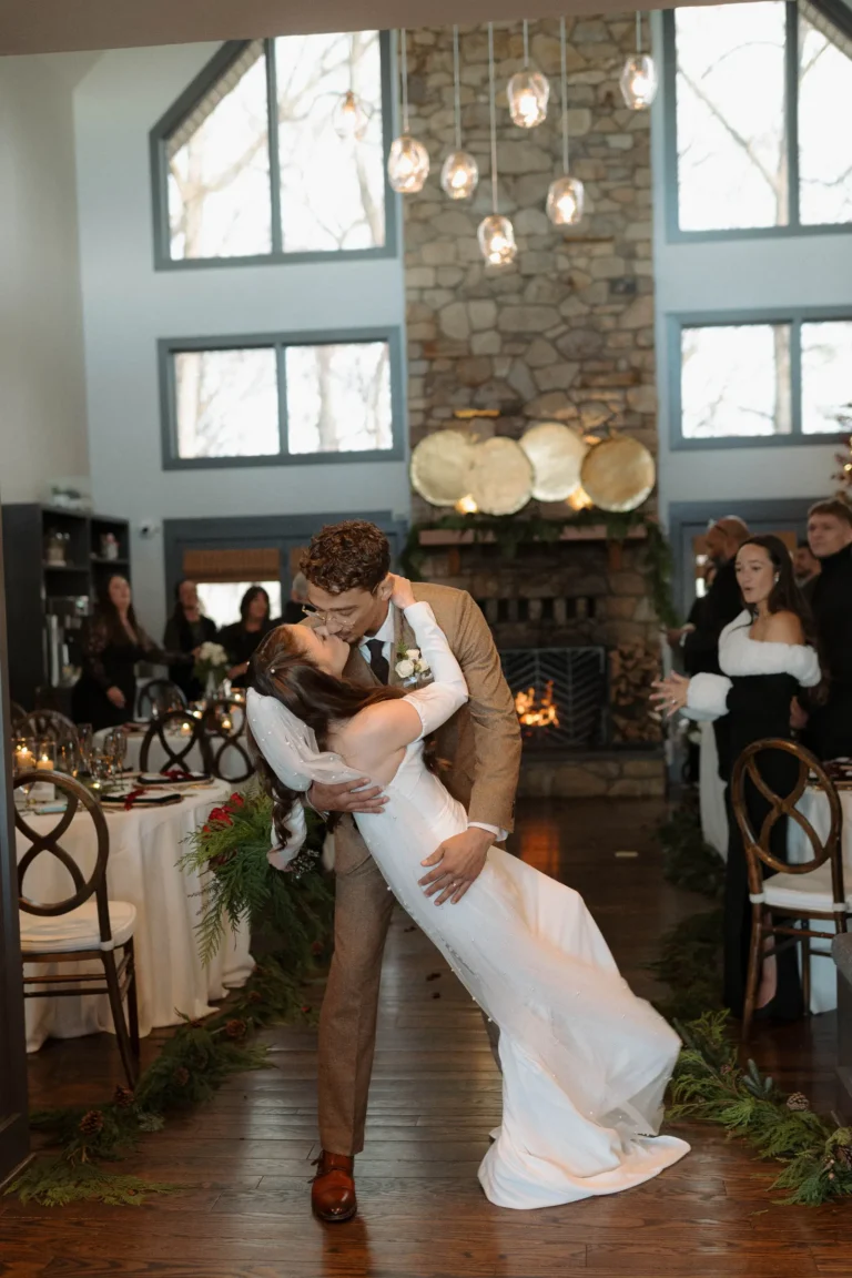 Groom dips his bride for a romantic kiss in a indoor reception hall lit by a stone fireplace.
