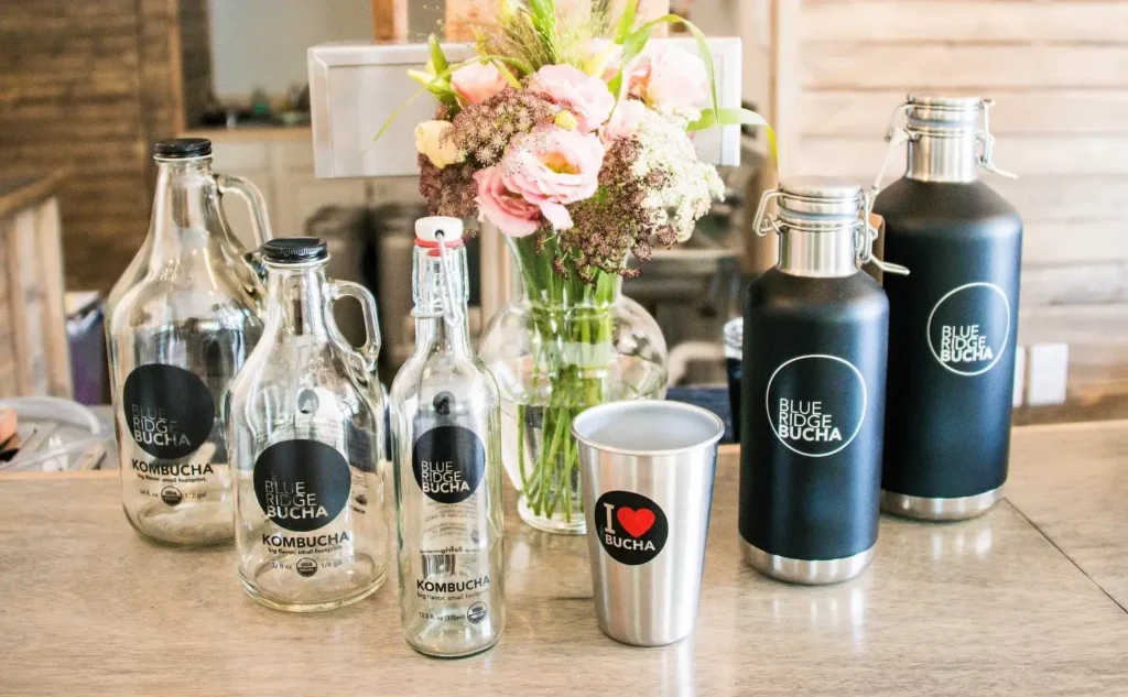 Blue Ridge Bucha kombucha bottles, growlers, and branded drinkware displayed on a wooden table with floral arrangement.